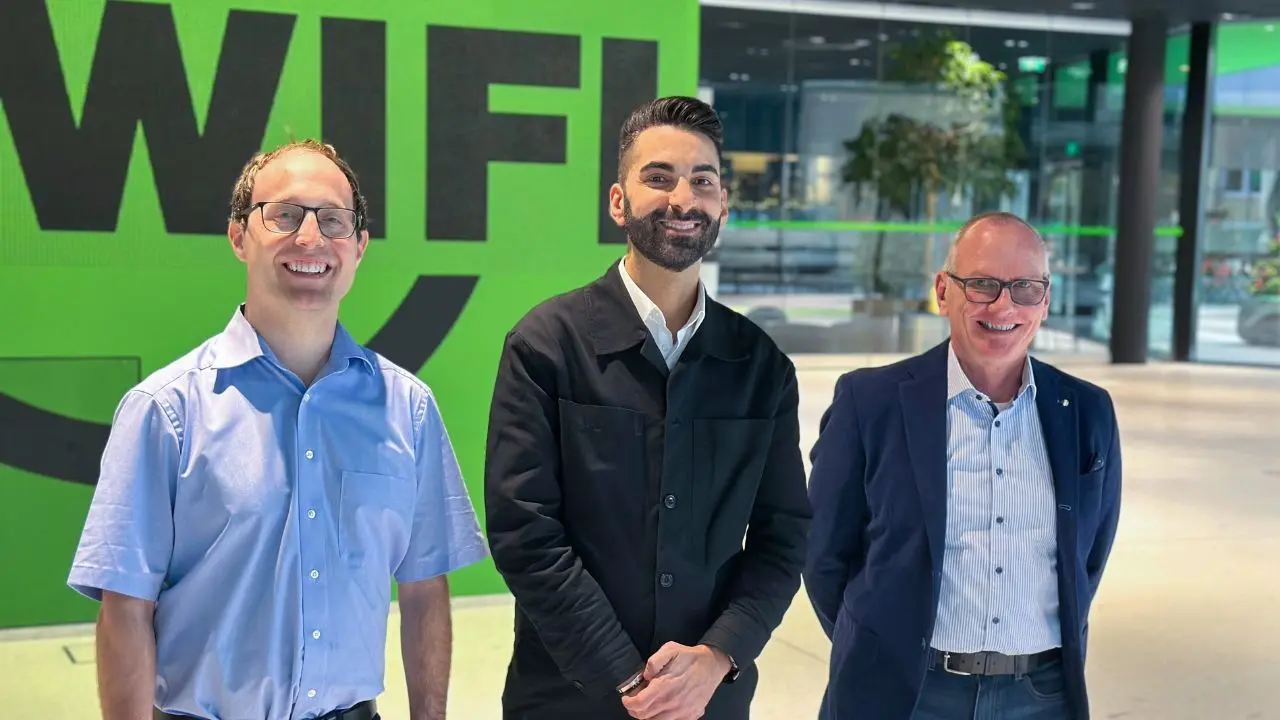Diese Grafik zeigt drei Männer in einer modernen Eingangshalle vor einer grünen Wand mit einem großen schwarzen Schriftzug "WIFI". Zwei tragen Hemden, einer eine Jacke. Sie stehen nebeneinander und blicken in Richtung Kamera.