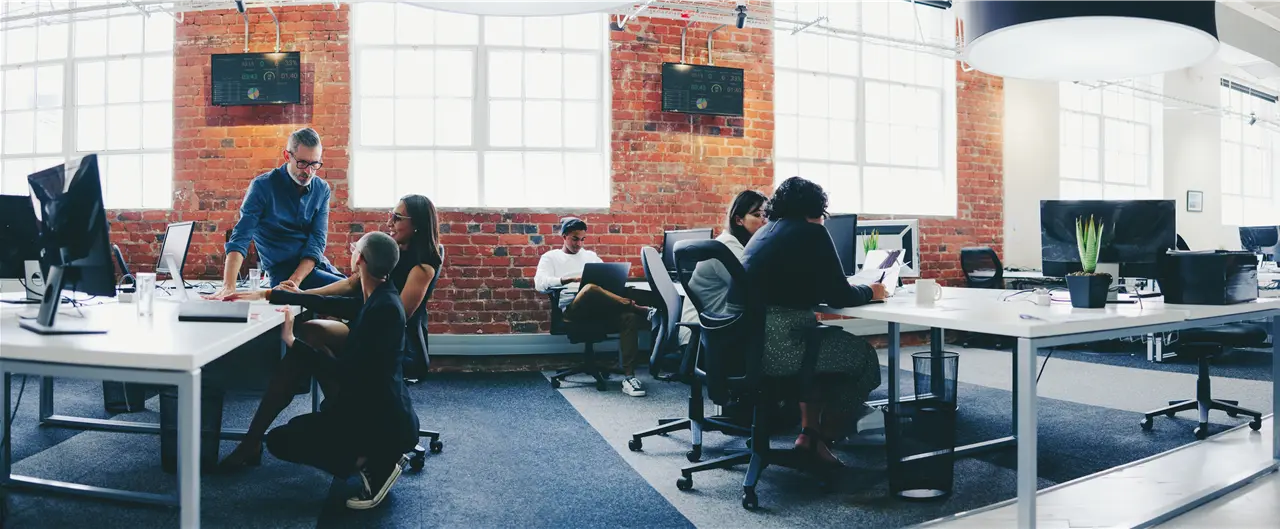 Menschen arbeiten in einem modernen B&uuml;ro mit Backsteinmauern, Computern und gro&szlig;en Fenstern. Manche diskutieren in Gruppen, andere arbeiten konzentriert an ihren Schreibtischen. Die Atmosph&auml;re wirkt energiegeladen und produktiv.
