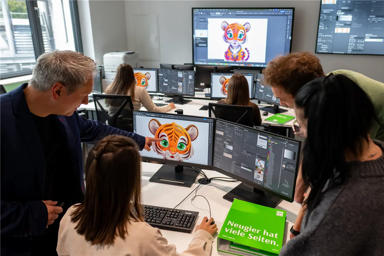 In einem modernen Büro arbeiten mehrere Personen an Computern, auf denen ein Tiger-Design erstellt und bearbeitet wird. Eine Gruppe bespricht die grafische Gestaltung. Ein großes Display zeigt das fertige Tiger-Bild.