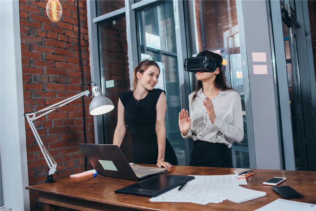 Zwei Personen in einem modernen B&uuml;ro: Eine Frau tr&auml;gt eine Virtual-Reality-Brille und gestikuliert, w&auml;hrend die andere sich anlehnt und den Computerbildschirm betrachtet. Davor liegen Papier und Laptop auf einem Holztisch.