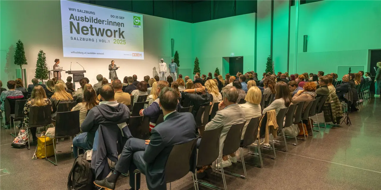 Eine Gruppe von Menschen sitzt in einem Konferenzraum; ein Sprecher steht auf der B&uuml;hne vor einer gro&szlig;en Leinwand; das Event ist mit "Ausbilder:innen Network Salzburg 2025" betitelt.