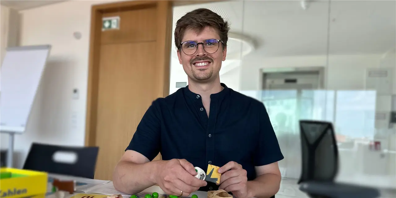 Ein Mann sitzt lächelnd an einem Tisch und spielt mit Holzspielzeug. Er trägt eine Brille und ein dunkelblaues Hemd. Der Hintergrund zeigt ein modernes Büro mit Flipchart und Stuhl.