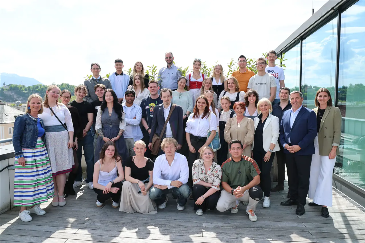 Eine Gruppe von Menschen unterschiedlichen Alters steht auf einer Terrasse mit Glasfront und Bergblick. Die meisten tragen legere Kleidung. Es scheint ein fröhliches Treffen oder Event zu sein. Die Umgebung ist sonnig und freundlich.