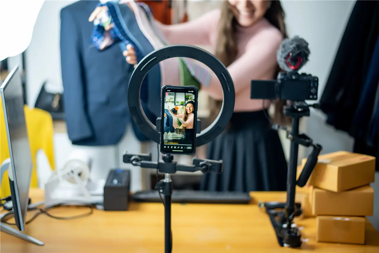Die Grafik zeigt eine junge Frau, die mit einem Smartphone und Ringlicht in einem Raum mit Kleidung auf einem Tisch ein Video aufnimmt. Eine zweite Kamera steht daneben. Dies wirkt wie die Aufnahme eines Modevideos oder Livestreams.