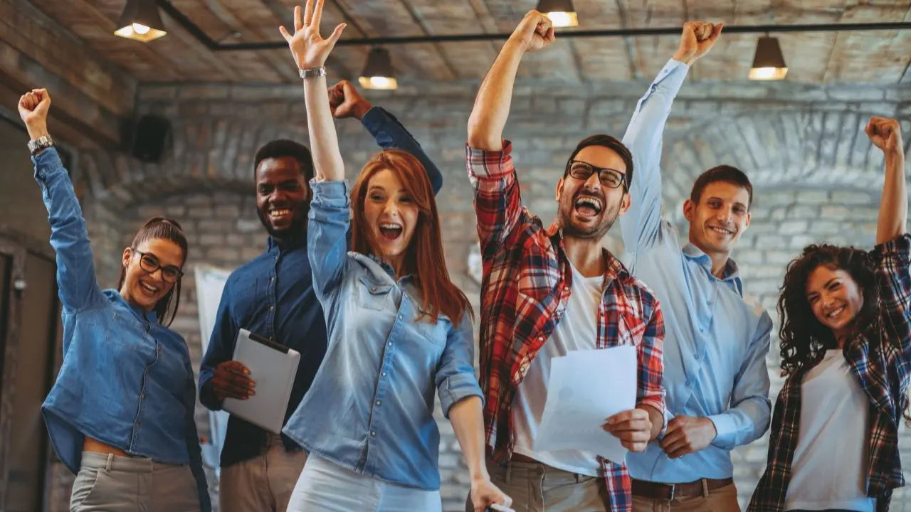 Eine Gruppe von sechs Menschen feiert mit erhobenen Fäusten in einer modernen Umgebung. Sie sehen glücklich und motiviert aus. Möglicherweise feiern sie einen Erfolg oder ein gemeinsames Ziel.