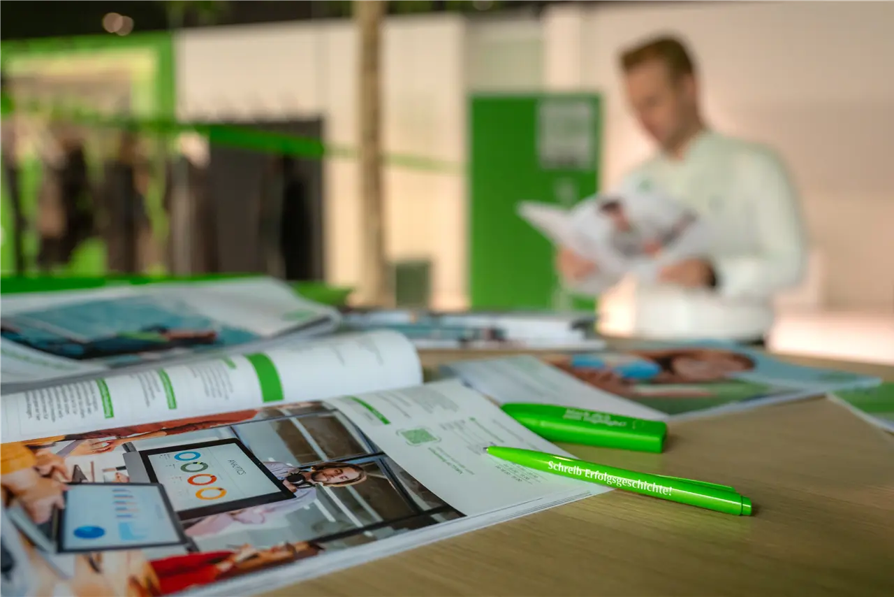 Diese Grafik zeigt einen Tisch mit Broschüren und grünen Stiften im Vordergrund, während im Hintergrund eine Person etwas liest. Die Szene wirkt geschäftlich und informativ.