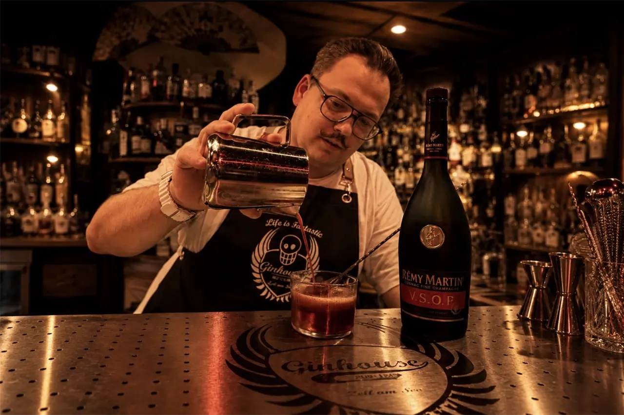 Ein Mann steht hinter einer Bar und gießt einen Drink aus einem Edelstahlbehälter ein. Neben ihm steht eine Flasche Rémy Martin Cognac. Die Bar ist gemütlich beleuchtet, voller Flaschen und Utensilien.