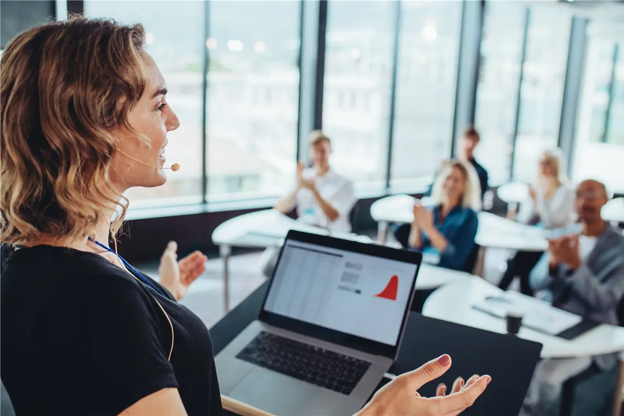 Eine Frau h&auml;lt eine Pr&auml;sentation vor einem kleinen Publikum in einem modernen B&uuml;ro. Auf ihrem Laptop sind Daten und ein Diagramm sichtbar. Die Zuh&ouml;rer sitzen an Tischen und klatschen.