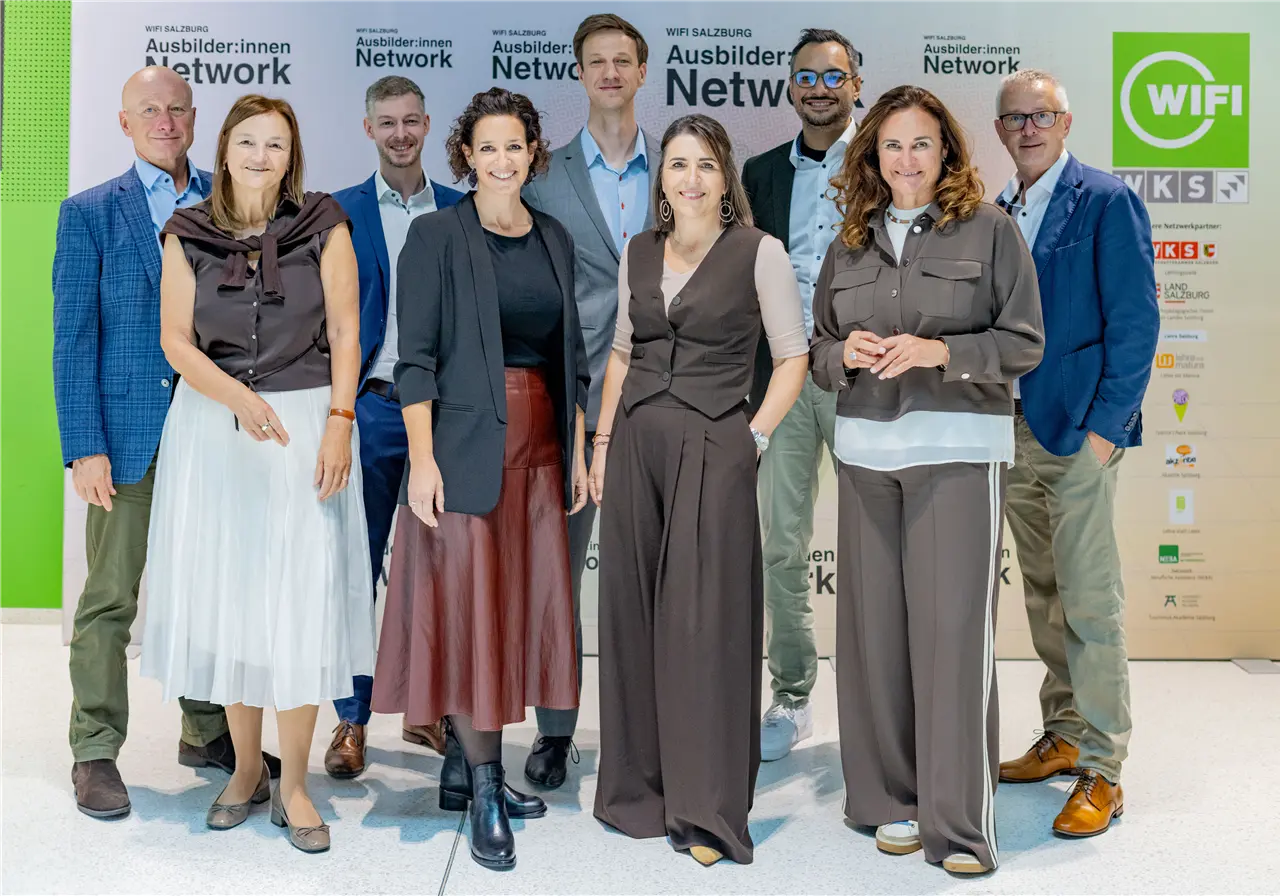 Eine Gruppe von neun Personen posiert lächelnd vor einem Hintergrund mit dem Logo "WIFI Salzburg Ausbilder:innen Network". Sie sind geschäftlich gekleidet und stehen vor einer Werbewand.
