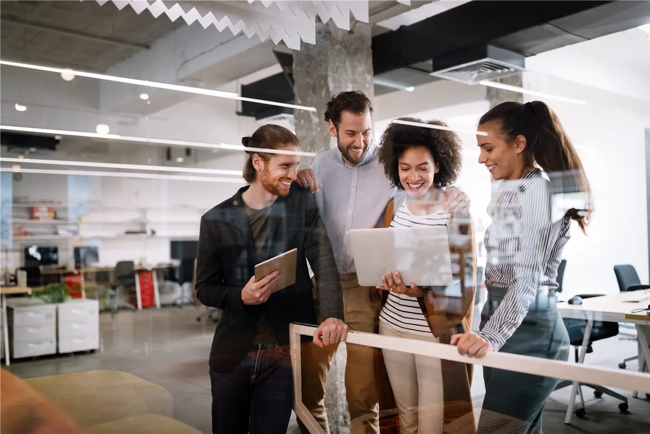 Dieses Bild zeigt eine Gruppe von vier Personen in einem modernen B&uuml;ro. Sie stehen zusammen und halten Tablets und Laptops, w&auml;hrend sie offenbar ein Gespr&auml;ch f&uuml;hren. Die Umgebung ist hell und offen gestaltet.