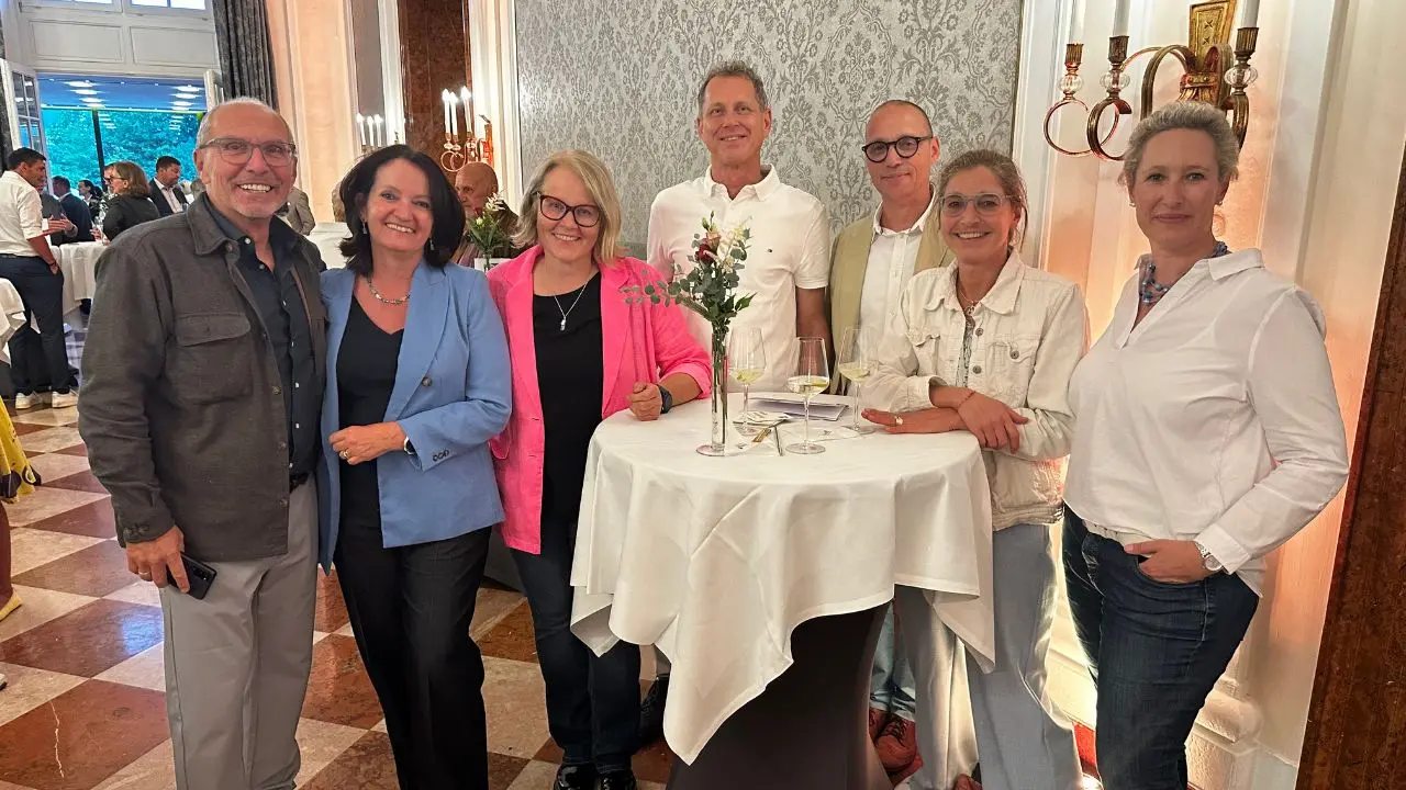 Eine Gruppe von sechs Personen steht um einen Tisch mit Blumen und Getränken. Sie sind in eleganter Kleidung und befinden sich in einem stilvollen Veranstaltungsraum mit Tapeten und Wandleuchtern.