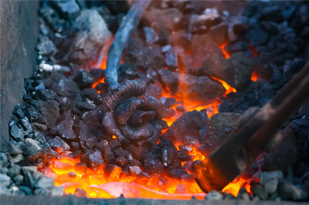 Das Bild zeigt einen gl&uuml;henden Metallgegenstand, der in einer Schmiede erhitzt wird. Kohlen und gl&uuml;hende Kohlenst&uuml;cke umgeben das metallische Objekt, das sich mitten im Herstellungsprozess befindet.