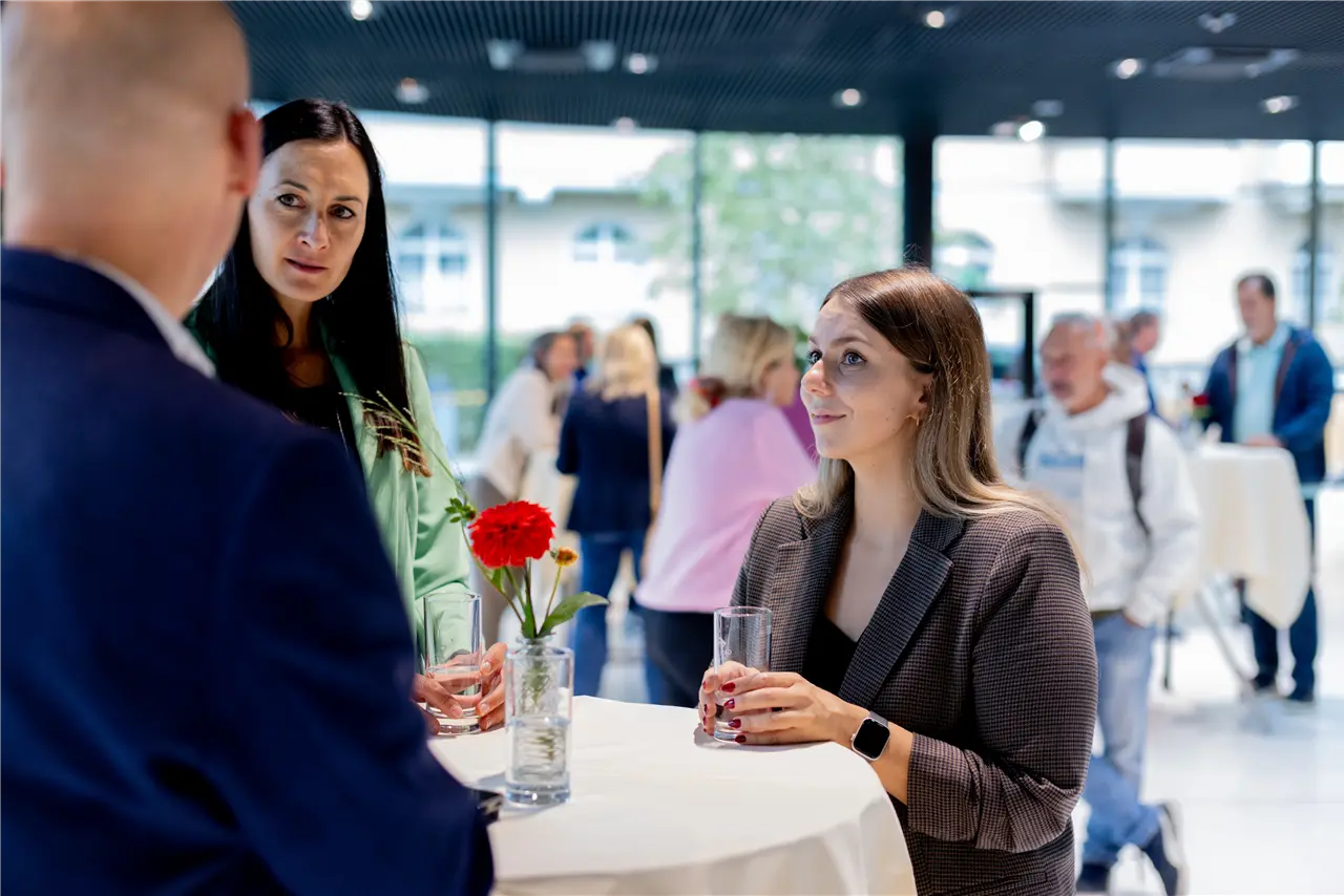 In einem gut beleuchteten Raum stehen mehrere Personen an einem Tisch, trinken ein Getränk und unterhalten sich, während im Hintergrund weitere Menschen zu sehen sind.
