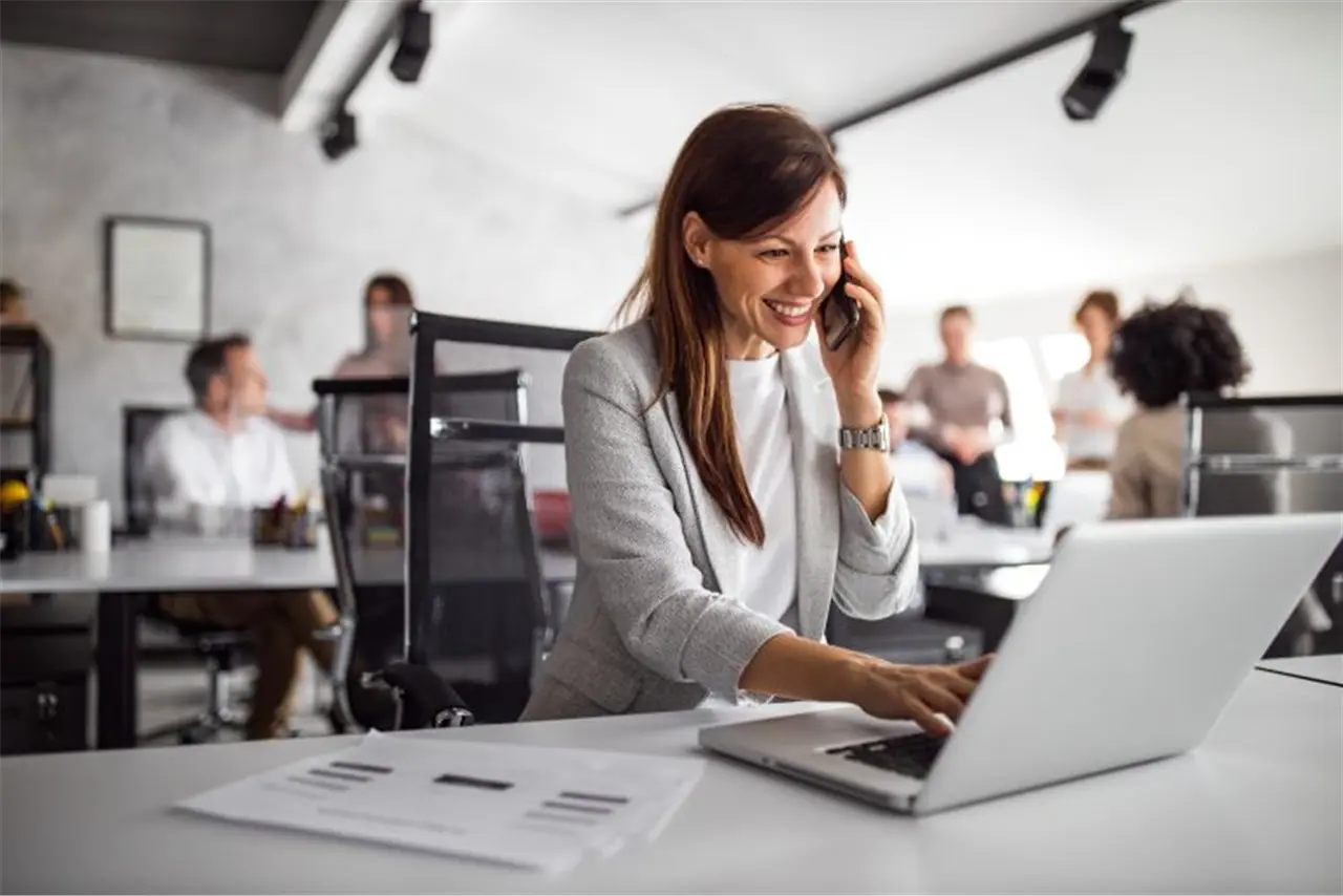 Eine Frau in einem modernen B&uuml;ro telefoniert und arbeitet am Laptop. Im Hintergrund sind mehrere Personen besch&auml;ftigt. Der Fokus liegt auf der professionellen und gesch&auml;ftigen Atmosph&auml;re.