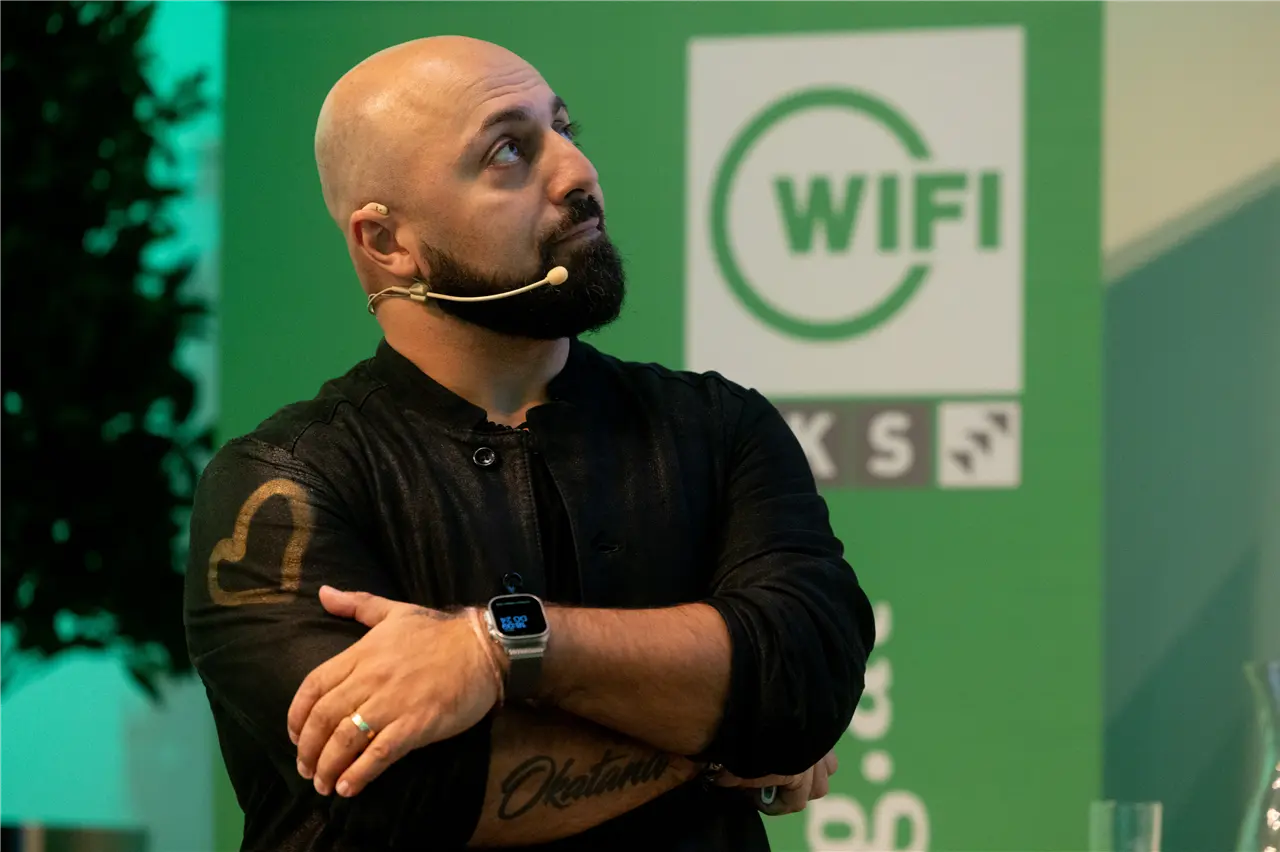 Ein Mann mit einem Headset steht vor einem grünen Hintergrund mit einem "WIFI" Logo. Er trägt schwarze Kleidung und eine Uhr, blickt nach oben und scheint sich auf eine Präsentation oder ein Event zu konzentrieren.