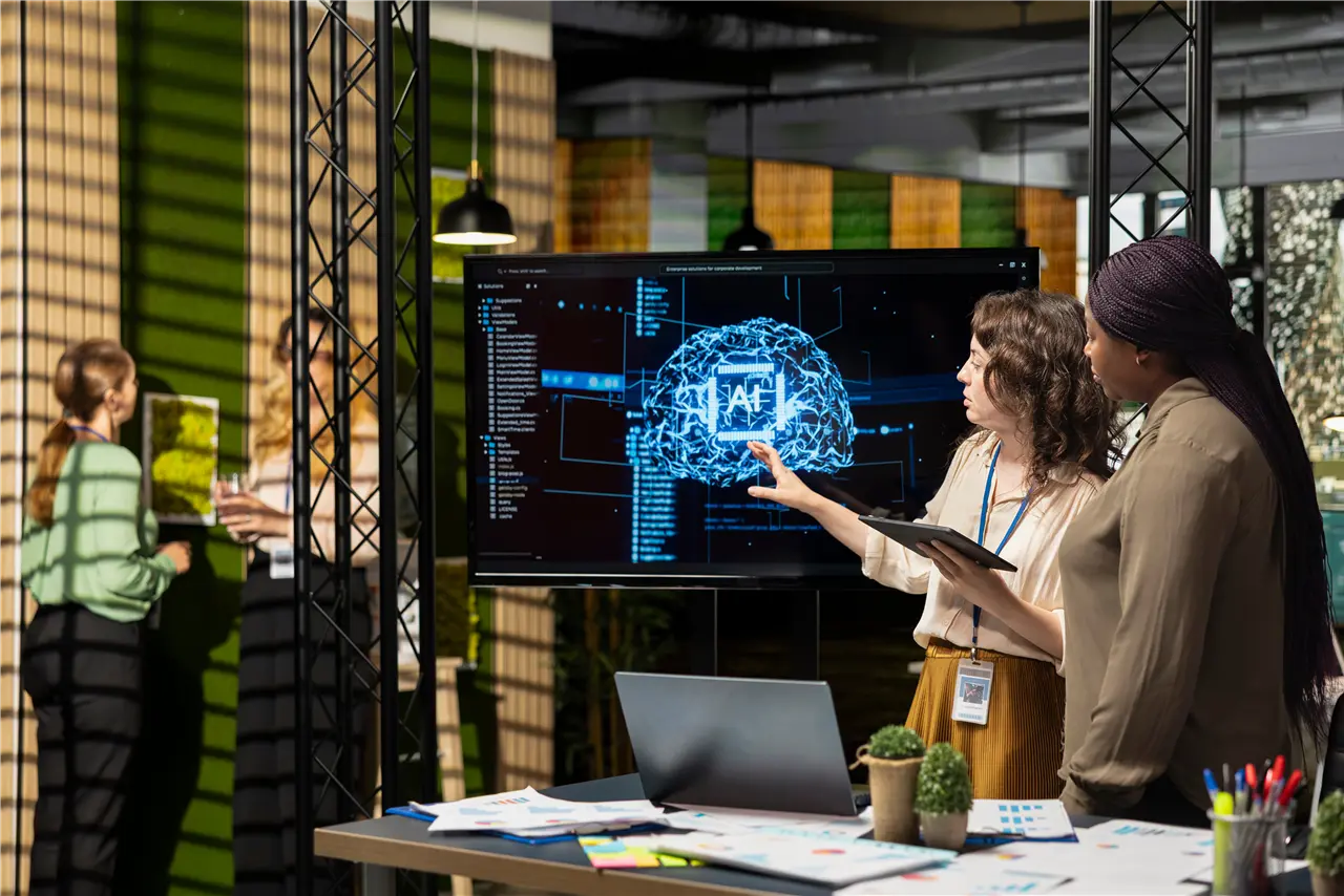 In einem modernen B&uuml;ro diskutieren zwei Personen anhand einer Bildschirmpr&auml;sentation &uuml;ber k&uuml;nstliche Intelligenz. Weitere Personen sind im Hintergrund.