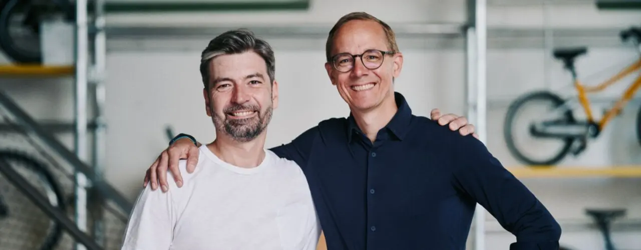 Zwei Personen stehen nebeneinander und lächeln in die Kamera. Eine Person trägt ein weißes T-Shirt, die andere ein dunkles Hemd. Im Hintergrund sind Fahrradteile sichtbar. Das Bild vermittelt Freundschaft und Zusammenarbeit.