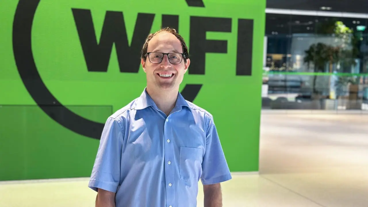 Die Grafik zeigt einen Mann in einem hellblauen Hemd vor einem großen grünen Schild mit der Aufschrift „WIFI“. Im Hintergrund sind moderne Gebäude mit Glasfassaden und Grünpflanzen zu sehen.