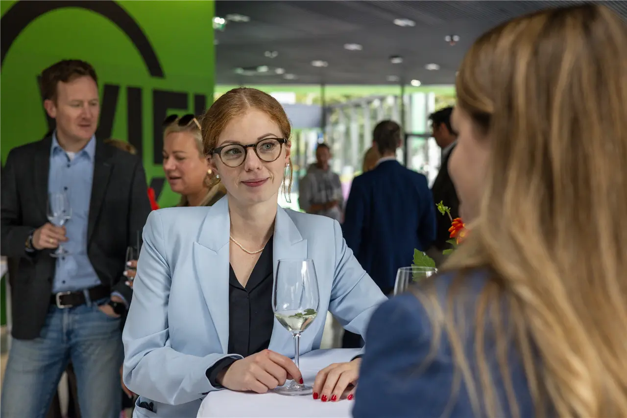Ein Gruppen von Menschen steht in einem modernen Raum zusammen und unterhält sich, während sie Wein trinken. Sie sind schick gekleidet und scheinen sich auf einer gesellschaftlichen Veranstaltung oder einem Empfang zu befinden.