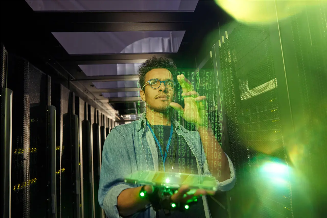 Eine Person interagiert in einem Serverraum mit einem holografischen Interface. Der Raum ist beleuchtet, und gr&uuml;ne digitale Zahlen scheinen auf dem holografischen Bild.